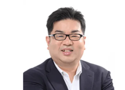 眼鏡をかけたアジア系の男性が、紺色のジャケットと白いシャツを着用し、白い背景の前で笑顔を見せている上半身のポートレート