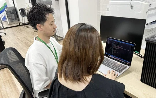 オフィスで男性と女性がノートパソコンを囲んで共同作業をしている様子。女性がパソコンを操作しており、画面にはプログラミングコードが表示されています。