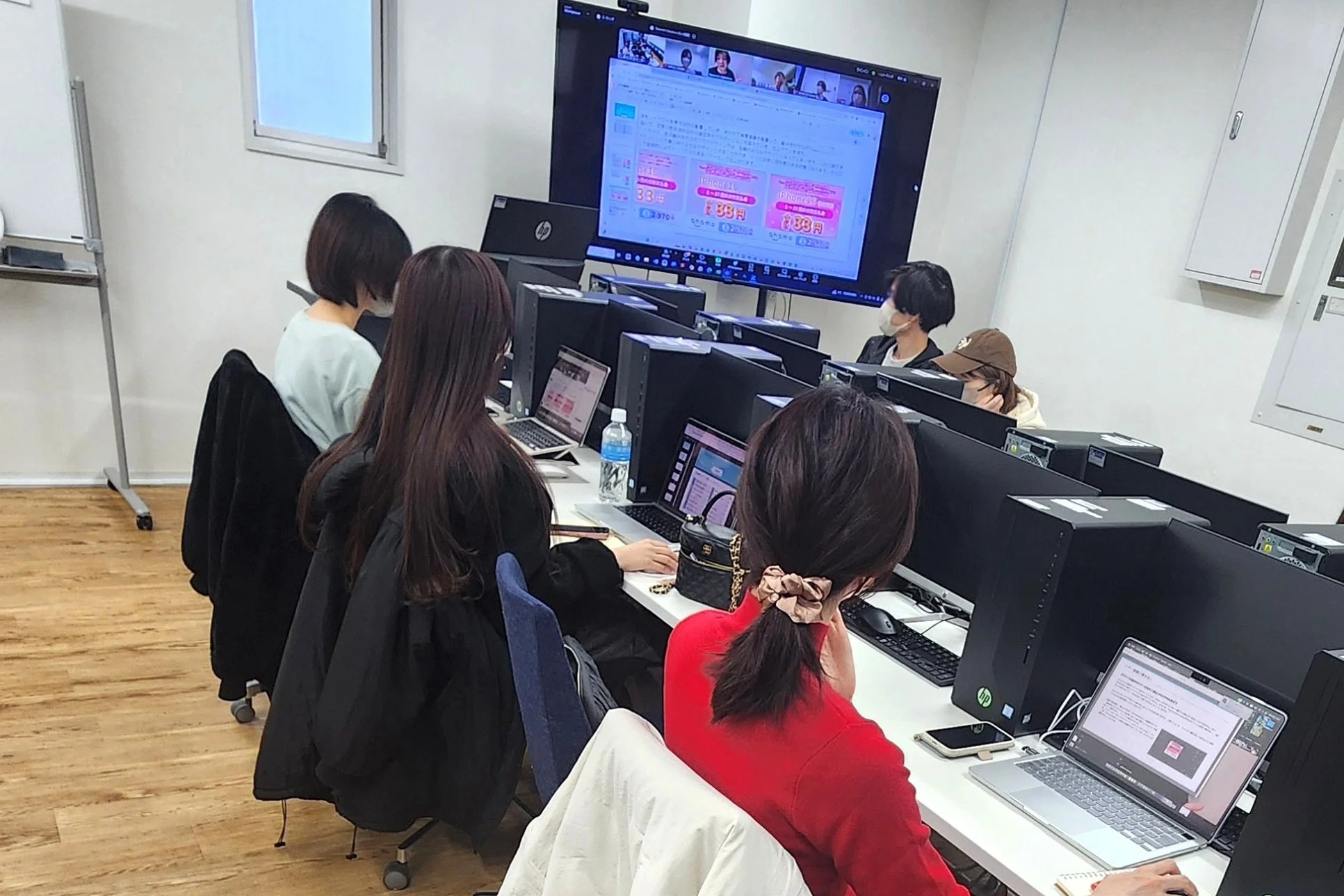 複数の人々が並んで座り、各自のラップトップや正面の大型モニターを見ながら作業している様子