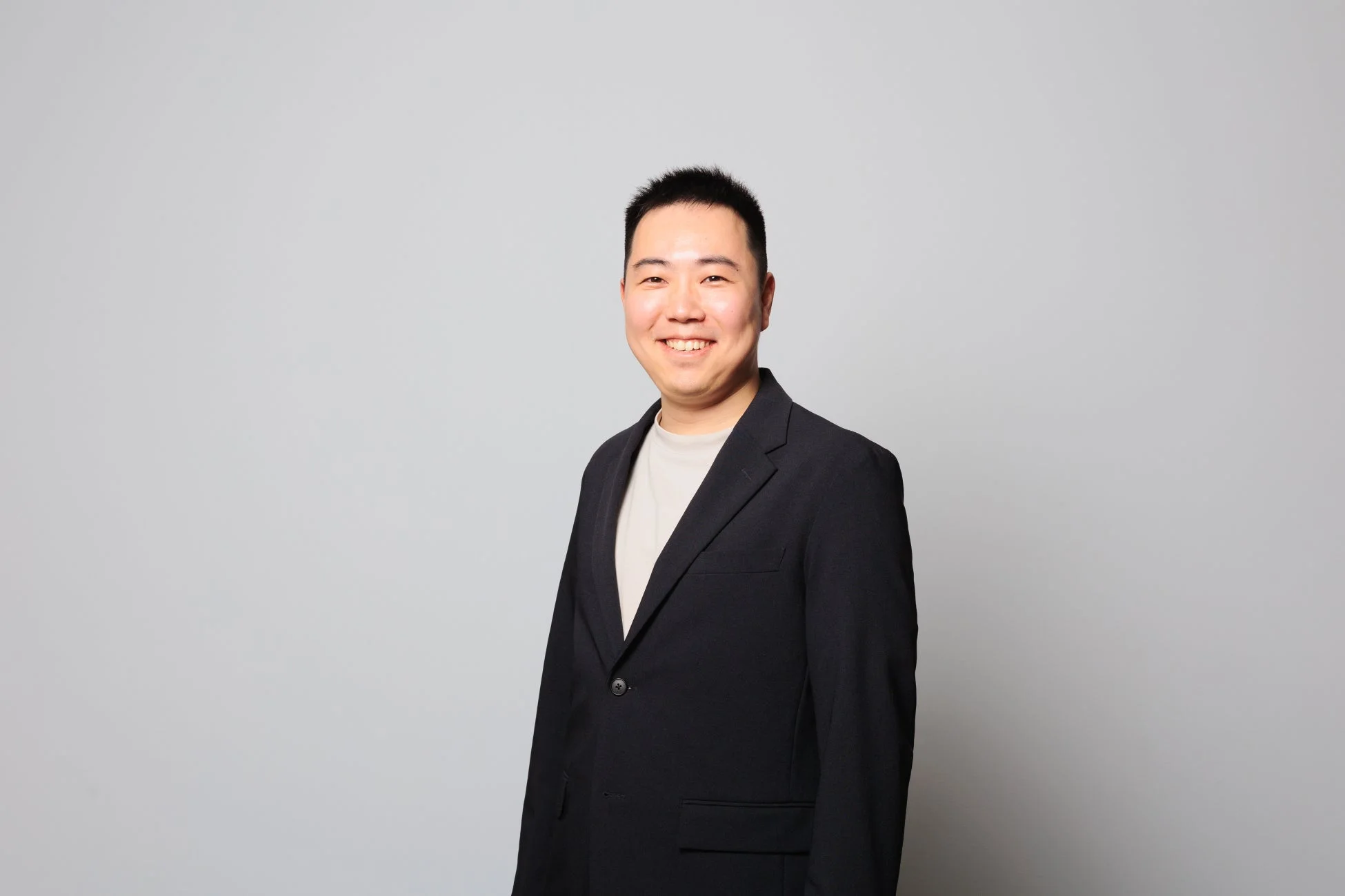 明いグレーの背景の前で、ダークスーツのジャケットとTシャツを着用したアジア系の男性が笑顔で立っているポートレート写真