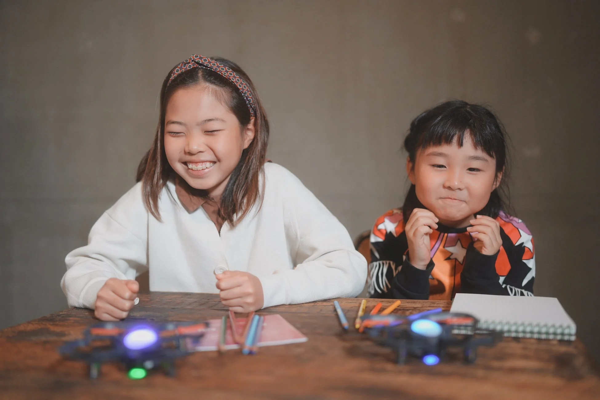 木製のテーブルに座る2人の女の子が、光る小さなドローンを囲んでいます。左の女の子は目を閉じて満面の笑みを浮かべ、右の女の子も楽しげな表情をしています。ドローンを使った学習や遊びの様子を捉えた、明るい雰囲気の画像です。