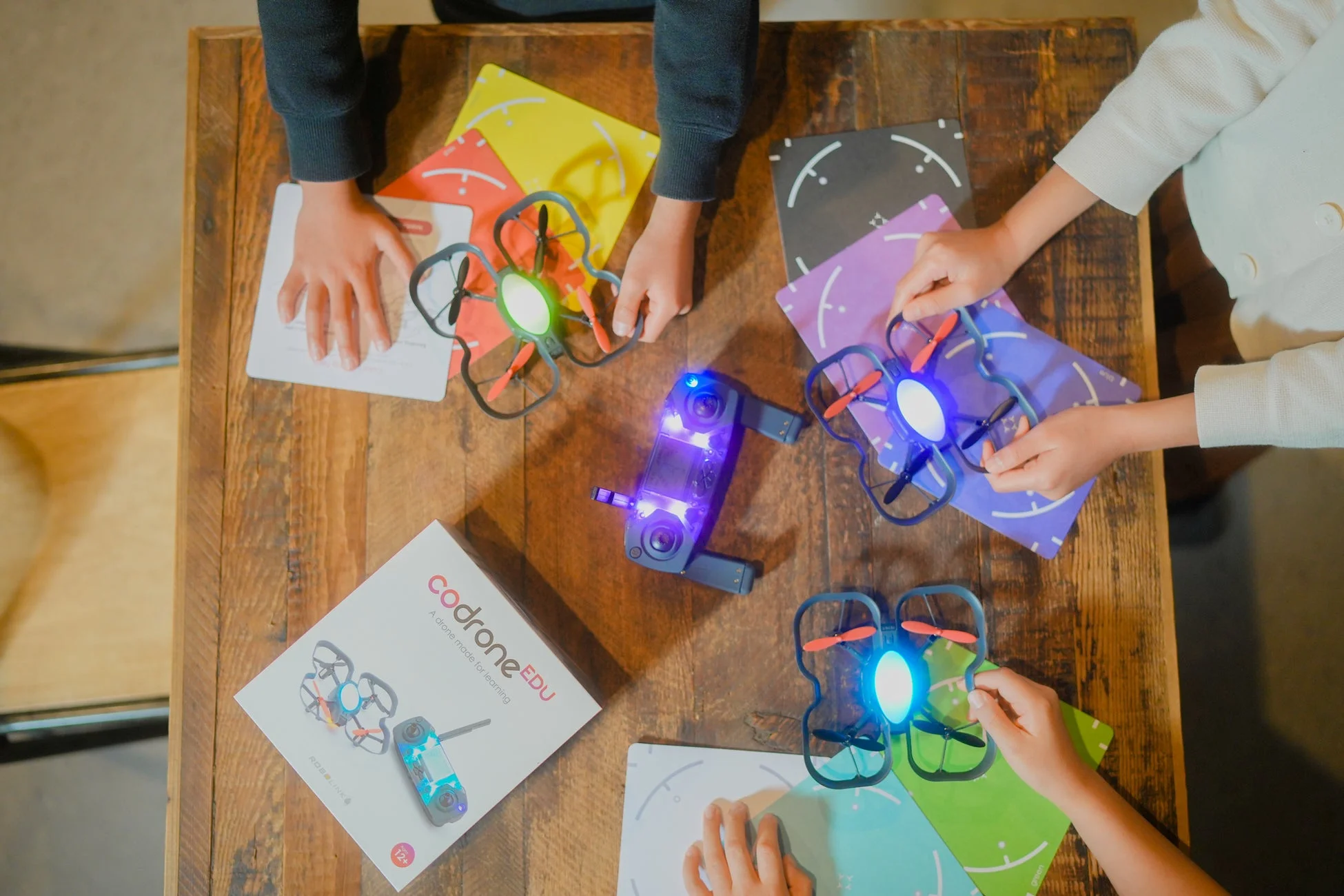 木製のテーブルを囲んで数人の子供たちが小型ドローンやコントローラーを操作している様子を上から捉えた画像です。「codroneEDU」の箱があり、ドローンを使ったプログラミング教育や STEM学習の場であることが伺えます。子供たちが楽しそうに学ぶ、実践的な教育風景です。