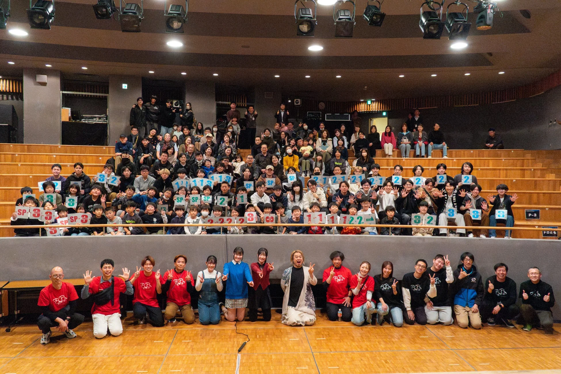 大勢の人々が劇場のような空間で撮影された集合写真です。多くの参加者が「SP」や数字の書かれたボードを持ち、楽しげな笑顔を見せています。