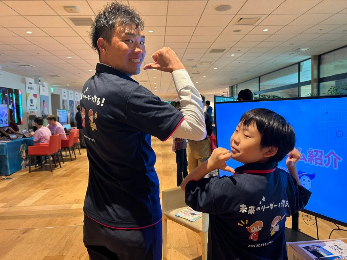 「未来のリーダーを作る！Kidsプログラミング」のTシャツを着た大人と子供が、イベント会場で笑顔でシャカサインをしています。子供向けプログラミング教室の活気ある様子が伝わる一枚です。