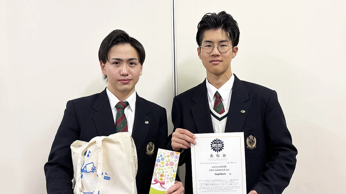 制服を着た2人の男子高校生が白い壁の前でポーズを取っています。右側の生徒は「U-18 SINGULARITY BATTLE QUEST 2023」の表彰状を持ち、左側の生徒は「learningBOX」と書かれたトートバッグを持っています
