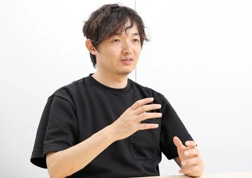 黒いTシャツを着たアジア人男性が、白い背景の前で座り、手を使って何かを説明している様子の画像