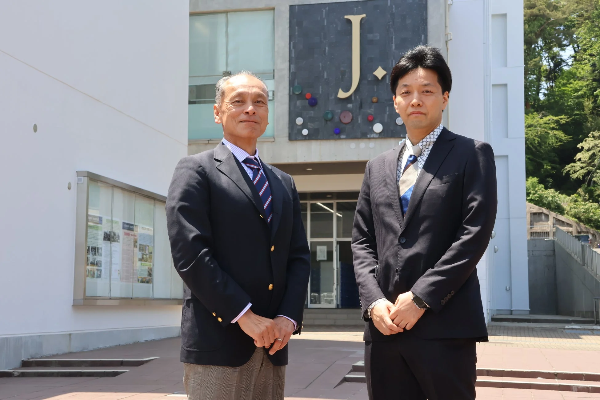 スーツ姿の男性二人とJ.のロゴがある建物