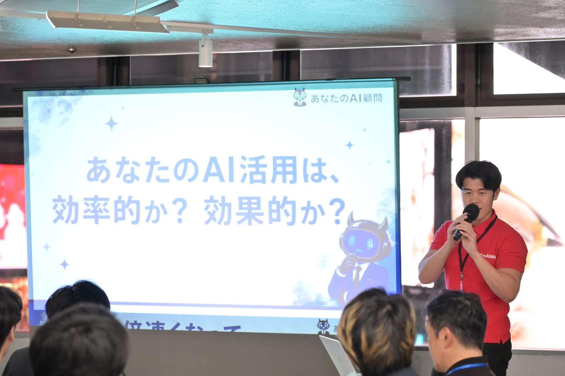 赤いシャツを着た男性がマイクを持ち、聴衆の前でプレゼンテーションを行っている