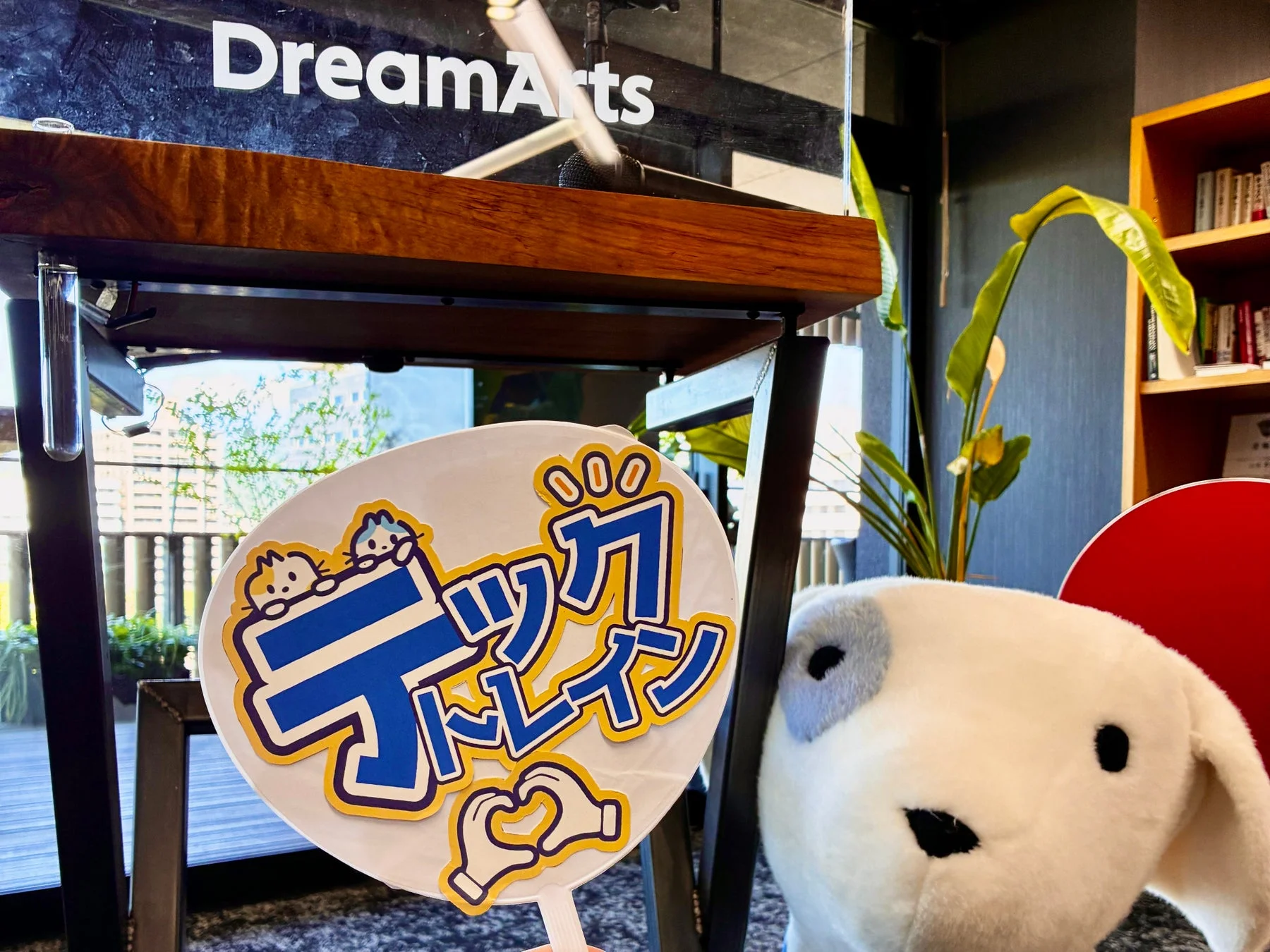 「DreamArts」のロゴが見える室内で、「ディックトレイン」のうちわと白い犬のぬいぐるみが手前に置かれている